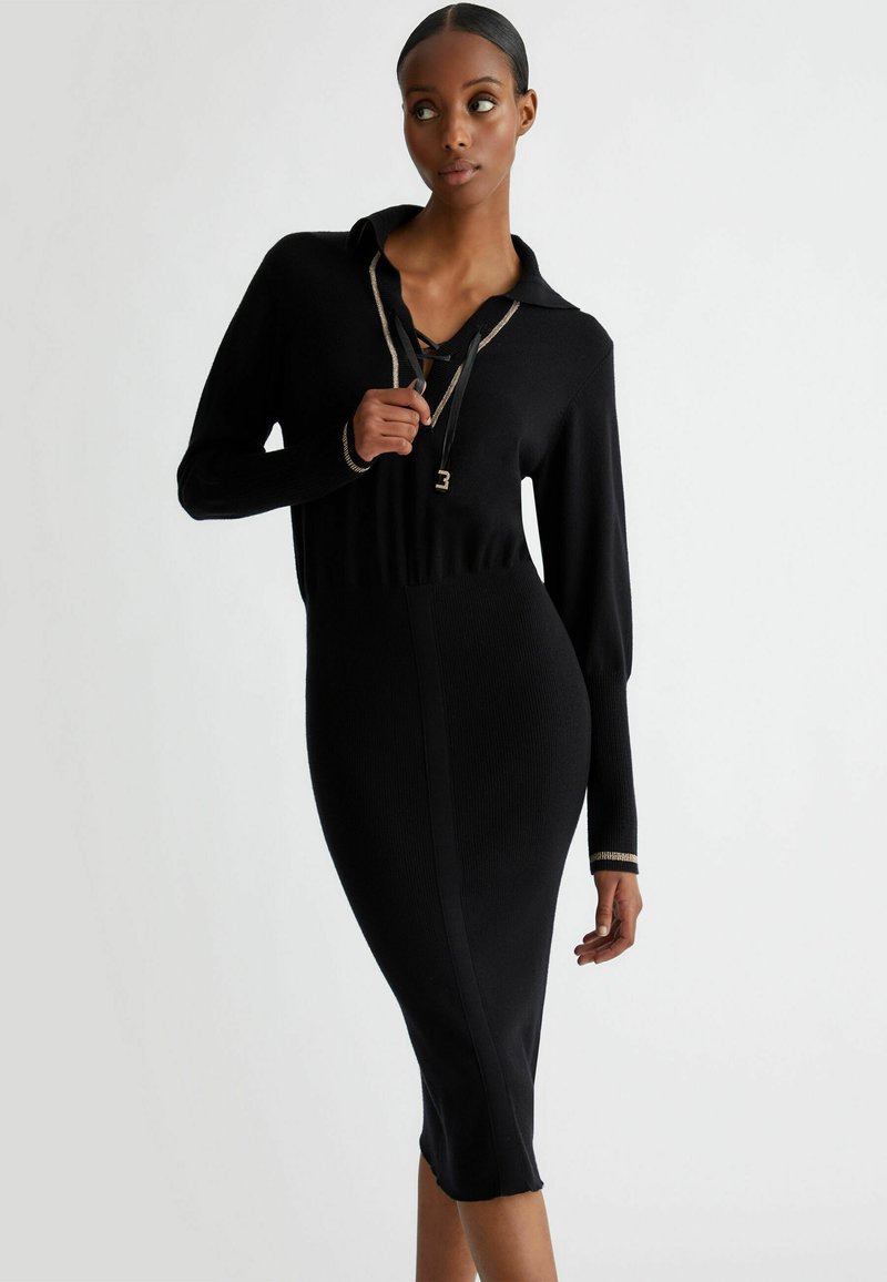 Zwarte gebreide jurk met een kraag, lange mouwen en een getailleerd silhouet. Opvallende details zijn een veterhals en gouden accentmanchetten.