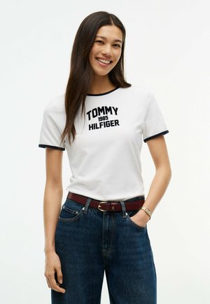 Femme souriante portant un t-shirt blanc Tommy Hilfiger avec bordure noire, jean bleu, ceinture marron et montre en or, debout devant un fond blanc.