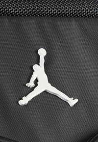 White Jumpman logo on a textured black fabric background. The logo depicts a figure leaping with a basketball.