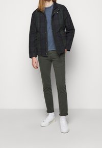 Donkergroene geruite jas met knopen, twee voorzakken, gedragen over een blauwe shirt, gecombineerd met olijfgroene broek en witte sneakers.