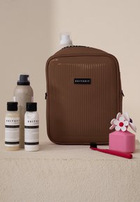 Braune, hartschalige Kulturtasche mit vertikaler Rillenstruktur, drei Flaschen mit Haarprodukten (weiß und beige), ein rosa blütenförmiger Behälter und eine rote Zahnbürste.