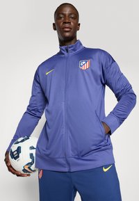 Ljubičasta sportska jakna sa zatvaračem koja prikazuje logo Atletico Madrida i žuti Nikeovo znak; model drži bijelu i plavu nogometnu loptu.