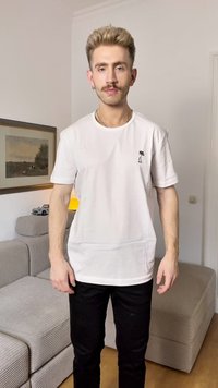 Weißes Baumwoll-T-Shirt mit kurzen Ärmeln, das ein kleines schwarzes Palmen-Graphic auf der Brust zeigt, kombiniert mit schwarzen Hosen, vor einem hellen Hintergrund.