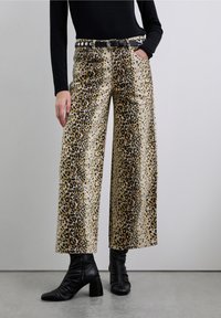 Pantalon large à imprimé léopard sur fond crème, avec une ceinture noire ornée d'une boucle en métal argenté. La texture est lisse avec des motifs distincts.