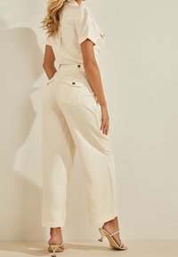 Kremový overal s krátkými rukávy, širokými nohavicemi a zadními kapsami. Má zlaté akcenty ve formě knoflíků a volnější střih. Jsou na něm nazouvací sandály na podpatku.