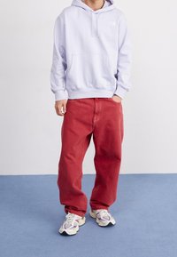 Sudadera con capucha de color púrpura claro y bolsillo frontal, combinada con vaqueros rojos holgados y zapatillas deportivas multicolores. Fondo liso.