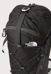 Crni ruksak ima teksturiranu tkaninu s mrežastim uzorkom, podesive naramenice i prednji džep s patentnim zatvaračem. Sadrži logo The North Face.