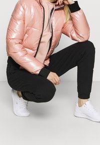 Rosa puffjacka med en glänsande finish, svarta detaljer och dragkedjestängning, matchad med svarta joggers och vita texturerade sneakers.