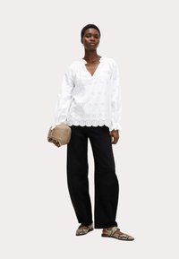 Blusa blanca con detalles de ojales, escote en V, mangas abullonadas. Combinada con pantalones amplios negros y un clutch beige. El calzado presenta un estampado de serpiente.