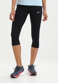 Femme portant des leggings crop noirs Nike et des chaussures de course bleu marine Nike avec des semelles blanches et rouges, se tenant sur un fond blanc.