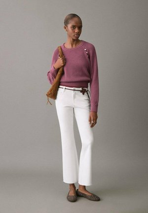 Femeie purtând un pulover tricotat mov cu nasturi, pantaloni evazați albi, scurtați, curea maro cu detaliu floral, ținând o geantă maro de umăr.