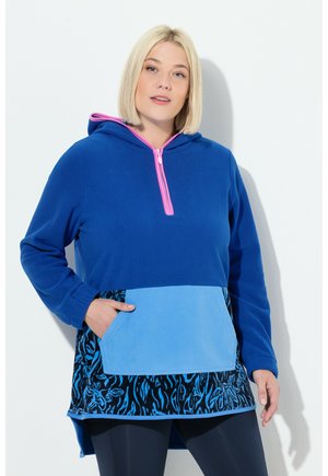 Frau trägt einen blauen Kapuzenpullover mit pinkem Reißverschluss, hellblauer Tasche und dunkelblauem Blumenmuster am Saum, steht mit einer Hand in der Tasche.