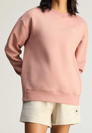 Osoba nosząca luźną różową bluzę i jasnobeżowe szorty z logo Adidas, stojąca z rękami za plecami na jednolitym tle.