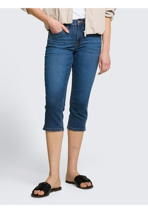 Frau trägt hochgekrempelte blaue Denim-Caprihose, beigen Blazer, weißes Oberteil und schwarze Slip-on-Sandalen, steht vor einem einfarbigen Hintergrund.