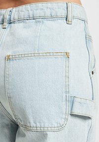Los pantalones de mezclilla de color azul claro presentan un bolsillo trasero con costura visible y una ligera textura, junto a un bolsillo lateral con detalle de solapa.