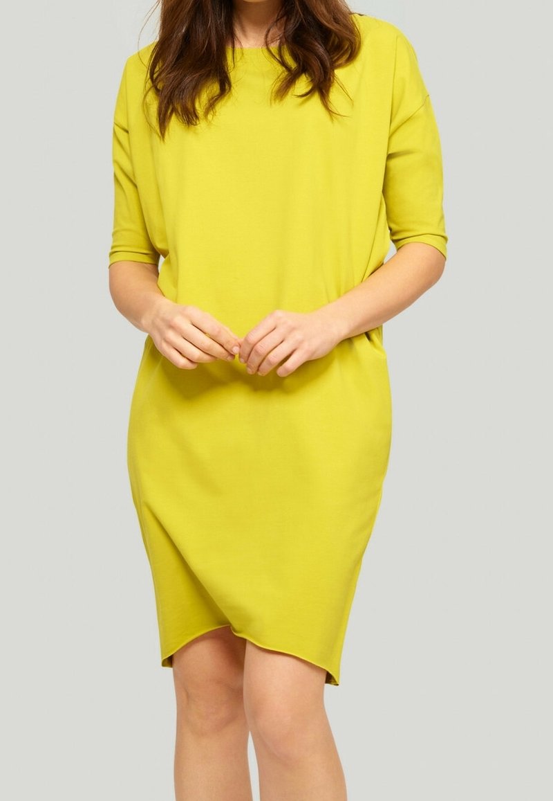 Femme portant une robe jaune vif longueur genou avec des manches trois-quarts, debout les mains jointes devant sur un fond uni.