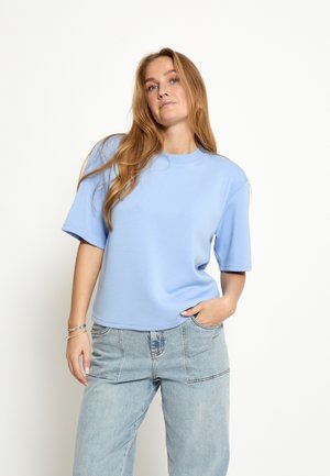 Jeune femme aux longs cheveux blonds portant un sweat-shirt à manches courtes bleu clair et un jean en denim bleu clair, debout devant un fond uni.