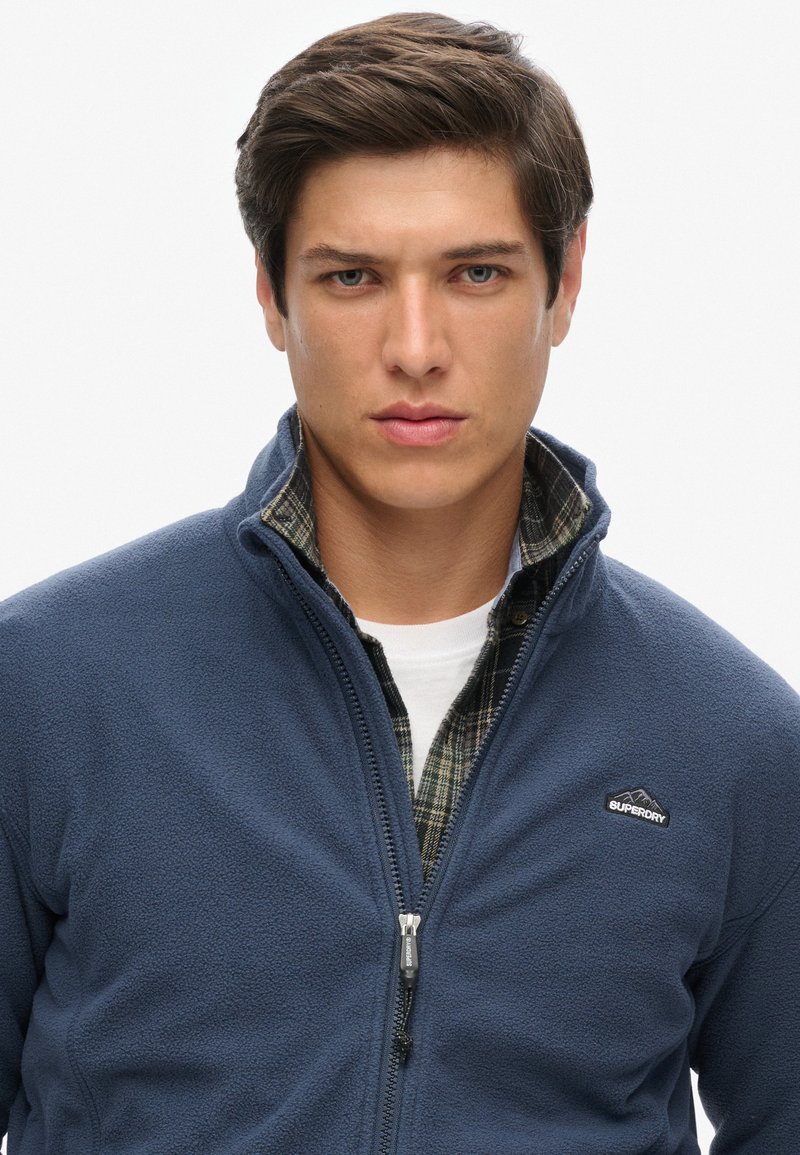 Granatowa polarowa kurtka z zamkiem, z kołnierzem stójkowym, z logo na lewej piersi, noszona na koszuli w kratę. Gładka tekstura.