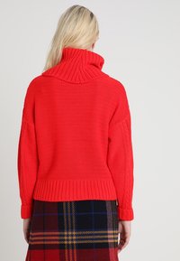 Červený pletený svetr s vysokým žebrovaným límcem, volným střihem a texturovaným vzorem. Nositelný přes kostkovanou sukni, který představuje útulný design.