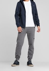 Hombre vistiendo una sudadera con cremallera azul marino, camiseta blanca, pantalones deportivos grises y zapatillas negras de pie frente a un fondo gris liso.