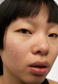 Gros plan sur un visage présentant une texture de peau lisse, des yeux marron clair et des lèvres légèrement teintées. Les cheveux sont raides et foncés avec une frange droite.