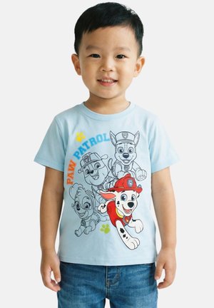 Jeune garçon souriant portant un t-shirt Paw Patrol bleu clair avec quatre personnages, dont un avec un casque de pompier rouge, et un jean bleu.