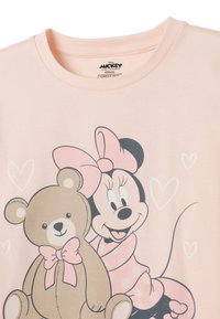 Camiseta de algodón rosa claro con un gráfico de un oso sonriente y un personaje con un lazo. Incluye corazones y lunares en la ropa.