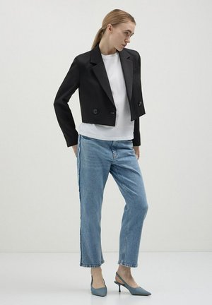 Veste noire cropped avec col cranté et double boutons, associée à une chemise blanche et un jean droit bleu clair, complétée par des talons bleus.