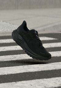 Μαύρο αθλητικό παπούτσι για τρέξιμο με παχιά σόλα, που αιωρείται πάνω από διάβαση πεζών σε δρόμο με άσφαλτο.