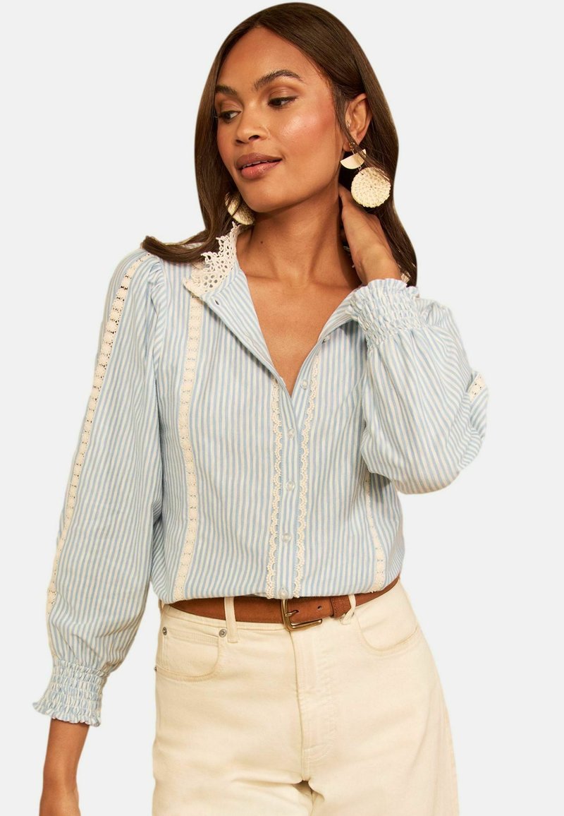 Woman wearing light blue striped blouse with lace details and puffed sleeves paired with beige pants and large round earrings.