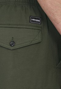 Khaki groene shorts van geweven stof, met een klepzak met een knop en een zwart label met de tekst "VOLCOM".