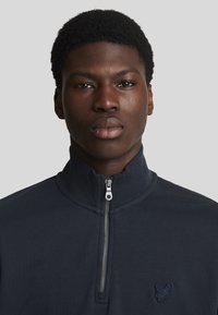 Marine blauwe zip-up sweatshirt met hoge kraag, gemaakt van zachte stof. Heeft een klein geborduurd logo op de borst en een zilveren ritsdetail.