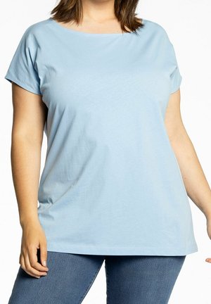 Kobieta w jasnoniebieskiej koszulce z krótkim rękawem i okrągłym dekoltem oraz niebieskich dżinsach, stojąca na białym tle.