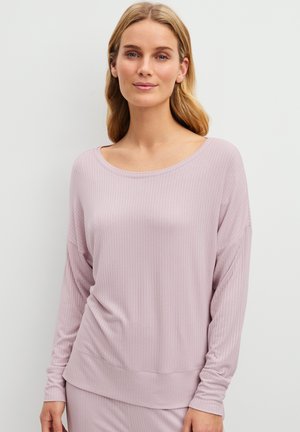 Vrouw met lichtbruin haar die een losse, lichtroze geribbelde top met lange mouwen en bijpassende broek draagt, staand tegen een effen achtergrond.