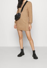 Beige Strickkleid mit gerippten Bündchen und Saum, kombiniert mit schwarzer Leder-Crossbody-Tasche und Stiefeletten mit dicken Sohlen.