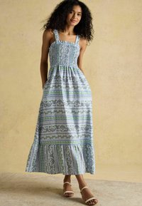 Maxi šaty v svetlomodrej farbe so zelenými kvetinovými vzormi, načechraný korzet, štvorcové ramienka a volánový lem. Nosené s bielymi sandálmi.