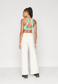 Αθλητικό σουτιέν σε ανοιχτό μπλε με σχεδιασμό σταυρωτής πλάτης και λεπτομέρεια από μεταλλικά στοιχεία, σε συνδυασμό με κρεμ παντελόνι φαρδιάς γραμμής, φορεμένο με μαύρα παπούτσια.