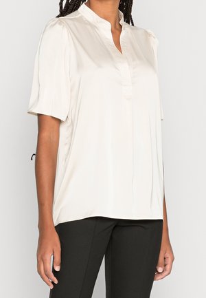 Femme portant un chemisier blanc à manches courtes avec un col droit et un pantalon noir, montrée du cou aux hanches sur un fond uni.