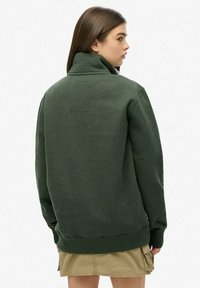 Sudadera de forro polar verde con cuello alto, puños y dobladillo acanalados. Presenta un diseño minimalista y un pequeño logo en la parte superior de la espalda.