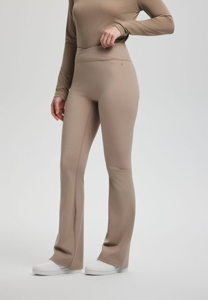 Persona con leggins acampanados beige y camiseta de manga larga, ajustándose la cintura, de pie sobre un fondo gris neutro con zapatillas blancas.