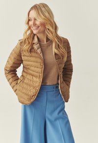 Doudoune marron clair avec coutures horizontales, dotée d'une fermeture à boutons et d'un col beige, associée à un pantalon bleu taille haute.