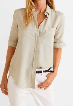Femme portant une chemise beige à boutons avec manches retroussées, un pantalon blanc et une ceinture noire tressée, debout avec une main dans la poche.