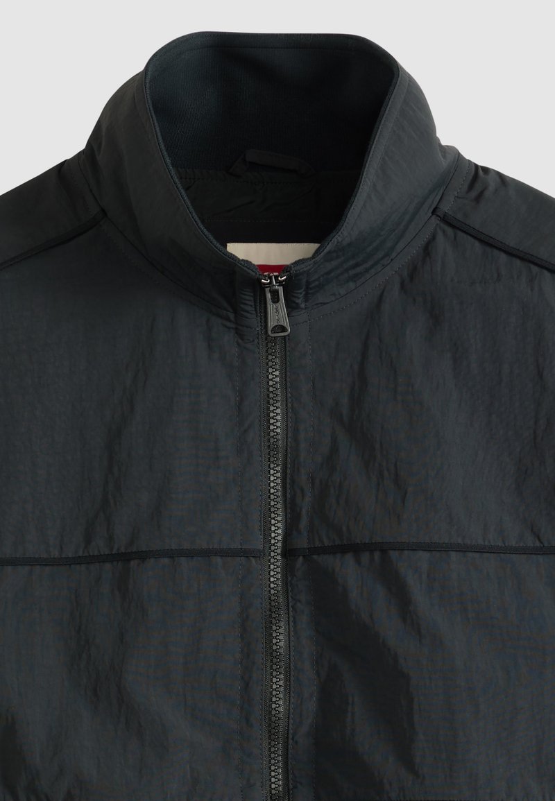 Zwarte lichte jas met hoge kraag en centrale ritssluiting, met zicht op het bovenste voorste gedeelte en de binnenkant van de kraag.