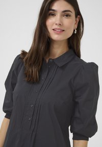 Mörkgrå blus med krage, plisserad front och puffärmar. Tillverkad av lättviktig tyg, med knappar längs fronten.