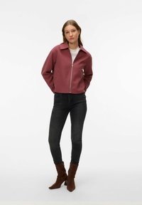 Femme portant une veste zippée rose poudré, un haut blanc, un jean skinny noir et des bottines à talons marron, debout les mains derrière le dos sur fond blanc.