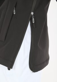 Veste softshell noire avec poches zippées, texture lisse et ourlet ajustable. Présente un marquage apparent sur les tirettes de zip.