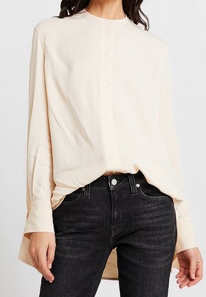Frau trägt ein cremefarbenes, langärmeliges Hemd mit Knöpfen, das in hoch taillierte schwarze Jeans gesteckt ist, und steht vor einem schlichten Hintergrund.