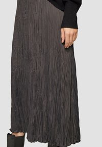 Jupe midi plissée en gris foncé, en tissu léger. Présente une surface texturée avec un ourlet volé. Portée avec un pull noir et des bottes.