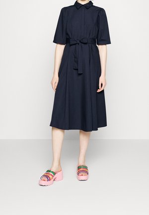 Kobieta ubrana w czarną sukienkę do kolan z wiązanym w pasie paskiem oraz różowe sandały na platformie z wielokolorowymi paskami.