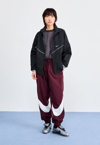 Svart vindjacka med vita accenter, randig grå topp, vinröda byxor med stort vitt swoosh-design, och grå sneakers med svarta detaljer.